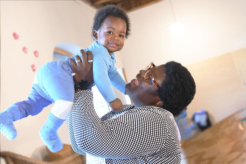 Eine Mutter hebt ihr Kleinkind hoch und schaut es liebevoll an – ein Moment voller Vertrauen und Nähe.